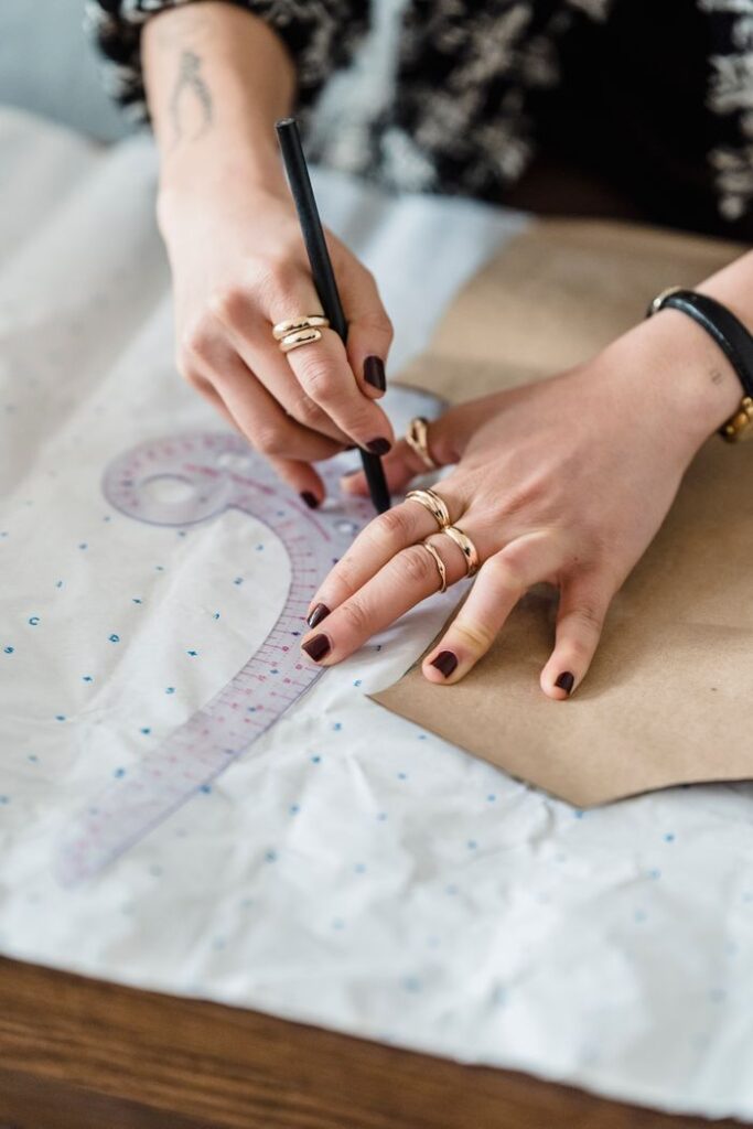 High angle of crop anonymous female dressmaker tracing with pencil on paper with carton template and curved ruler in workspace